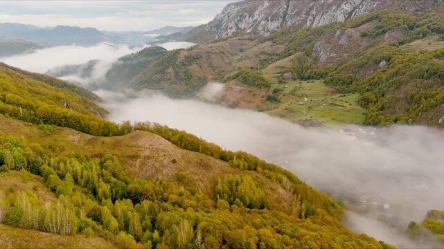 Aerial view of thick fog rolling through a valley between forested hills and rocky mountain peaks during autumn in Salciua de Jos, Alba County, Romania.