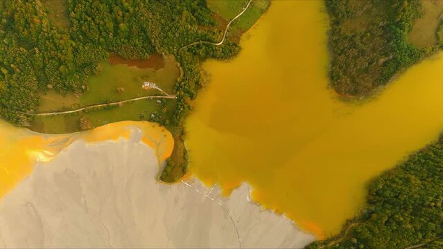 Aerial view of the toxic orange and grey tailings pond covering the sunken village of Geamana, surrounded by green forests in Geamana, Alba County, Romania.