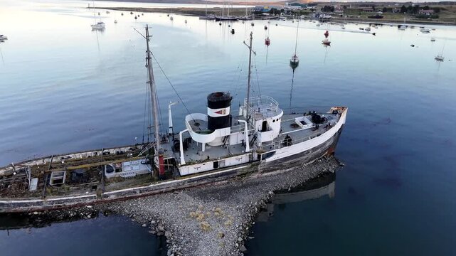 Cinematic drone lift revealing the historic Saint Christopher (HMS Justice) shipwreck grounded in the calm, reflective waters of Ushuaia harbor at dusk