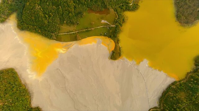 Aerial view of the toxic tailings pond with vibrant yellow and grey sludge surrounded by green forests at Geamana, Alba County, Romania.
