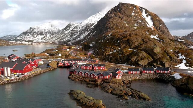 Aerial view of traditional red fishing cabins nestled along the rocky coastline with majestic snow-capped mountains and a calm bay in Nordland, Norway.