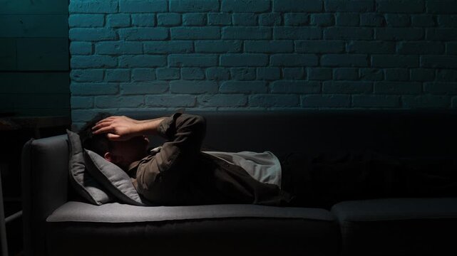 Man lying on a sofa in a dim brick walled room, covering his face with his hands, overwhelmed by insomnia, headache and emotional pain, isolated and exhausted at night