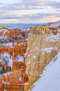 A dramatic close-up view of a snow-capped hoodoo spire against a backdrop of orange rock formations under a soft winter sky in Bryce Canyon.