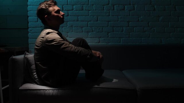 Man experiencing sadness and deep despair. Sitting hunched and withdrawn on a sofa in a dimly lit room. Creating a somber silhouette against a brick wall. Symbolizing mental health issues and solitude