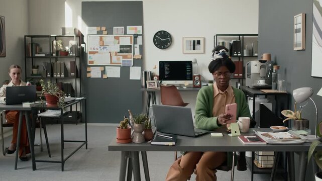 High angle shot of African American woman in vibrant clothes sitting down at her table and typing on laptop and using smartphone in moderm openspace office