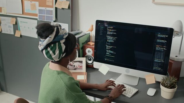High angle handheld zoom in shot of African American woman coding on computer at cluttered desk in modern office