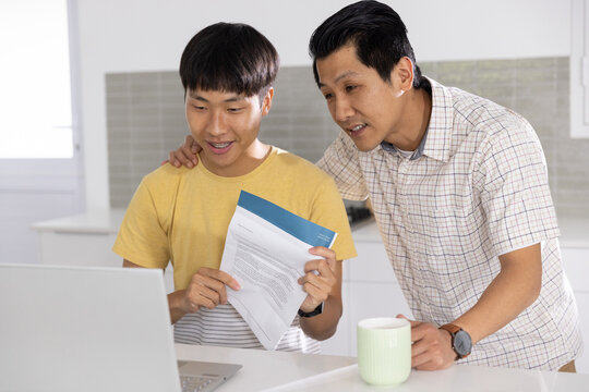 Two Asian males reviewing laptop and blue documents at kitchen, yellow tee near bright window