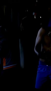 Vertical video: Training shirtless man adjusting gloves while teen in hoodie striking bag in garage