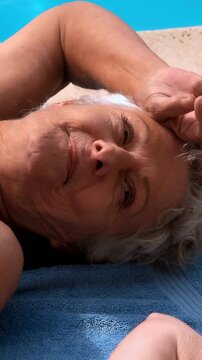 Vertical video: Lying older woman doing facial and hand moves by pool, forearm entering, strap seen