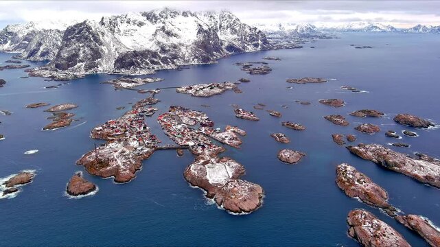 Aerial view of Henningsvaer fishing village on small islands surrounded by blue sea and snowy mountains in Nordland, Norway.
