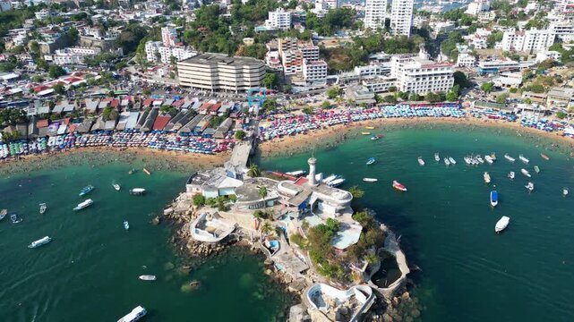 Aerial drone view of vibrant Caleta and Caletilla beaches, Acapulco, Mexico. Crowds enjoy sun, sand, clear water. Boats, city, green hills form a popular vacation scene.