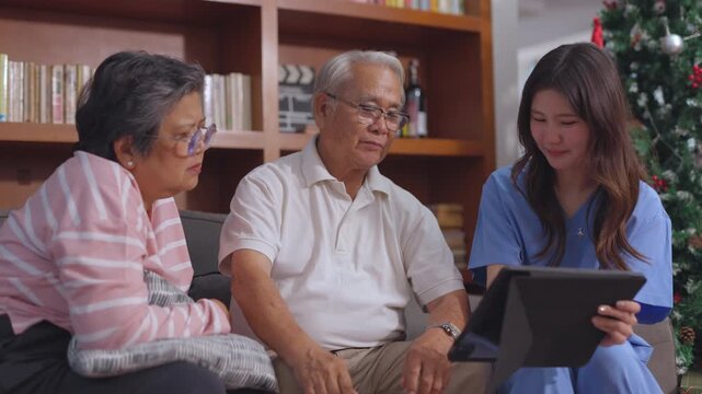Caring Asian caregiver and nurse explaining healthcare options on digital tablet to an elderly senior couple, showing medical results during happy consultation at home
