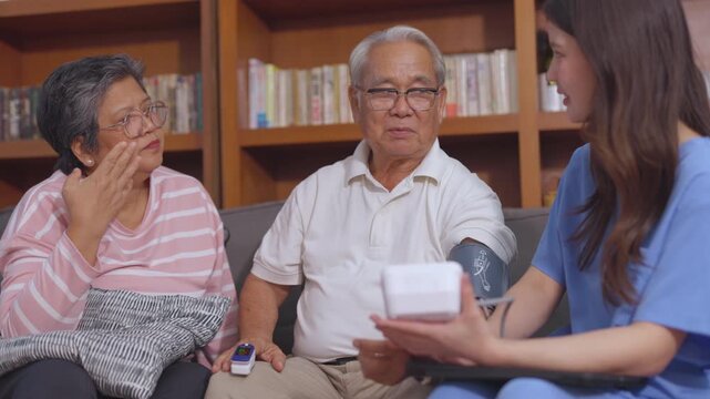 Attentive asian nurse, also caregiver, checks blood pressure for senior man, patient and husband. His wife, concerned woman, watches during home care visit with couple