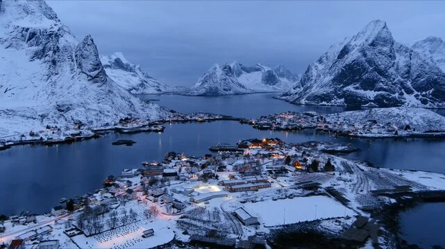 Aerial view of the snow-covered village of Reine in the Lofoten Islands with glowing house lights and dramatic mountains at twilight Nordland, Norway.