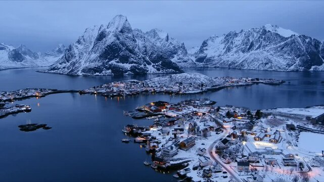 Aerial view of a snowy fishing village with illuminated houses nestled among dramatic mountains and calm fjords during blue hour in Nordland, Norway.