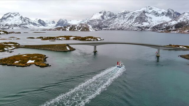 Aerial view of the Fredvang Bridges as a fishing boat leaves a wake in the water surrounded by snow-capped mountains in Nordland, Norway.