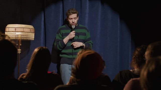 Charismatic young man performing stand up comedy monologue on stage and interacting with audience during open mic night