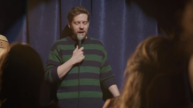 Young male comic holding microphone as performing stand up comedy routine on stage in front of laughing audience during open mic night at club