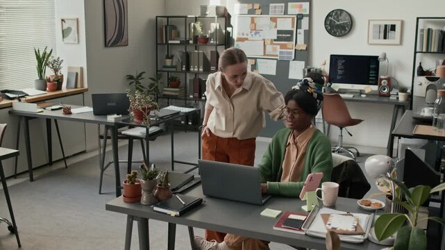 High angle shot of two female colleagues discussing work at laptop in a bright, modern office with several workstations
