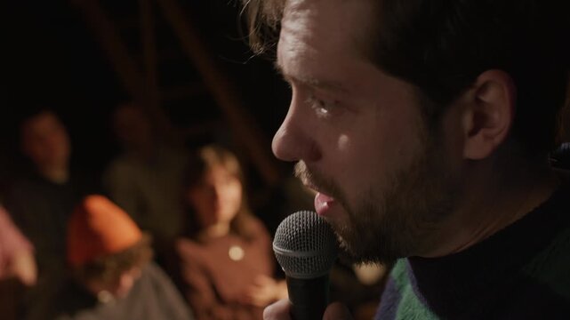 Young male comedian speaking into microphone and entertaining audience during live stand up show in in cozy dimly lit venue. Close up view