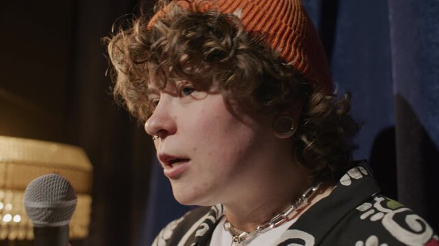 Curly-haired girl in beanie speaking into microphone while delivering stand up speech on comedy club stage. Close up view