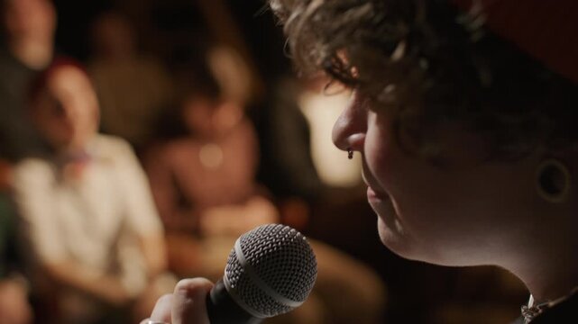Young female comedian performing live set of standup comedy in dark club, speaking to laughing audience at open mic night. Close up view