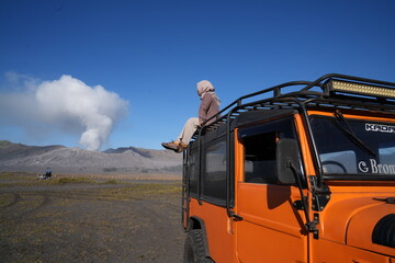 Young woman sitting on top of orange off-road jeep looking at Mount Bromo volcano. © demilayer