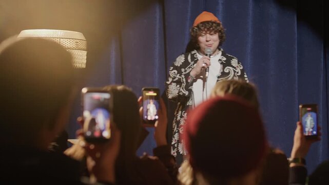 Young female comedian walking on stage of comedy club and performing stand up set with microphone as fans taking videos of her with mobile phones