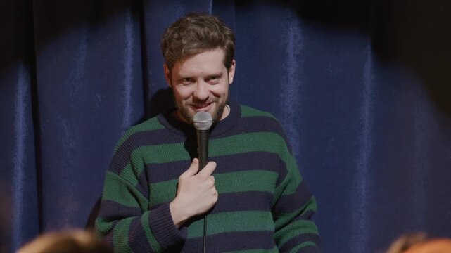 Young smiling male comic holding microphone, telling jokes and interacting with audience during live stand up set on comedy club stage