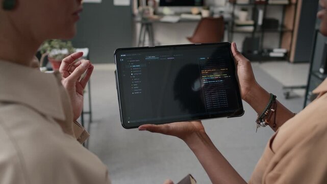 Low angle close up over the shoulder shot of two unrecognizable women in beige shirts, discussing program code displayed on screen of dark digital tablet in office