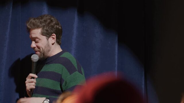 Male comedian standing against blue velvet backdrop, delivering standup speech with microphone during live event at comedy club