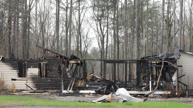 Burned house stands broken and blackened. Trees lean behind, silent and stark. Charred beams crumble in the wind. No people visible, no signs of life. Tragic aftermath of a devastating fire