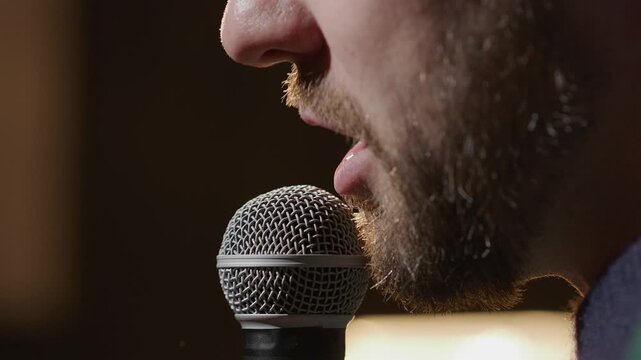 Extreme close up view of face of young bearded man speaking into professional microphone on stage during live performance or stand up show