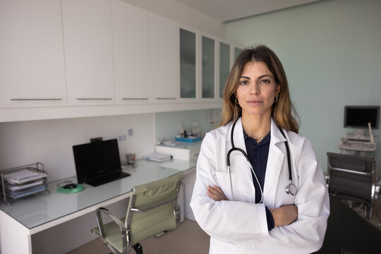 Portrait of Hispanic female doctor in white lab coat and stethoscope standing confidently with crossed arms in clinic office, looking at camera, exuding professionalism and readiness to provide care