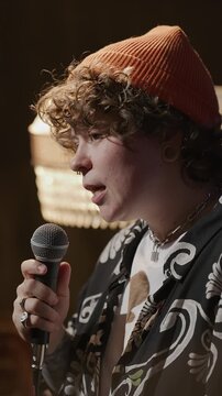 Young female Gen Z performer with curly hair and orange beanie speaking into microphone during emotional standup comedy show on stage. Vertical clip