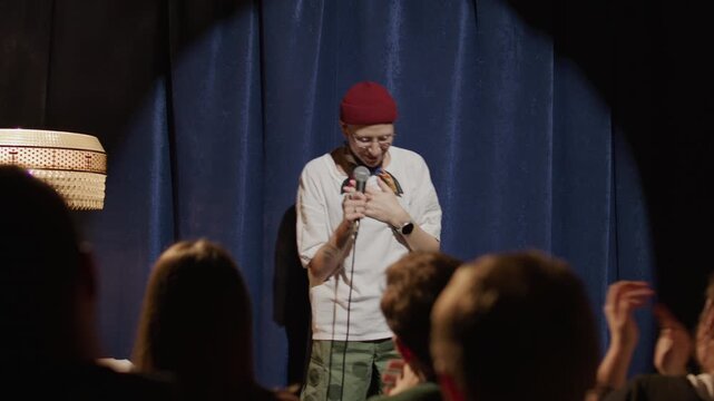 Young cheerful woman in trendy outfit waving to applauding audience, smiling and delivering stand up performance on stage of comedy club