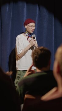 Smiling female comedian holding microphone as performing stand up show on stage, audience clapping and laughing during her monologue. Vertical format footage