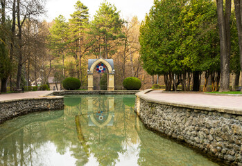 Naklejka premium View of Mirror Pond and the Glass Stream pavilion with its stained-glass window in the distance in Kislovodsk Lower Park at sunset in spring. Healthful walks at resort city park. Scenic landscape