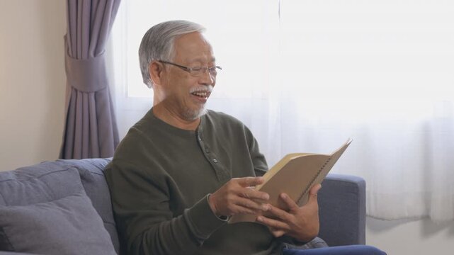 Peaceful senior Asian man at home, embracing quiet lifestyle. He finds joy reading book and reminiscing with vdo album, creating calm relax session on sofa