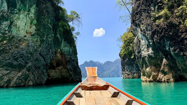 The wooden bow of a longtail boat sails between Khao Sam Kloe cliffs on Cheow Lan Lake in Khao Sok, point-of-view shot of moving towards famous landmark limestone rock towers in Thailand