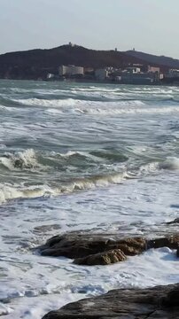 A flock of seagulls flies over crashing ocean waves on a sandy beach with modern city skyscrapers in the background in Dalian China.