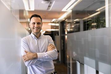 Fototapeta premium Portrait of confident business man or employee stands with arms crossed in office corridor, smiling, looking at camera, represents career growth and professional achievements. Corporate member profile