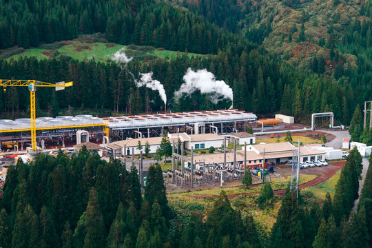 Aerial view of Ribeira Grande Geothermal Power Station with steam rising from cooling towers, surrounded by lush green forest and rolling hills in Ribeira Grande, Acores, Portugal.
