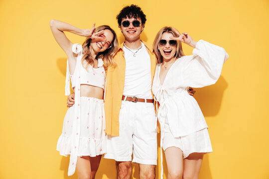 Group of young three stylish friends posing in studio. Fashion man and two cute female dressed in casual summer clothes. Smiling models having fun. Cheerful women and guy near yellow wall, sunglasses