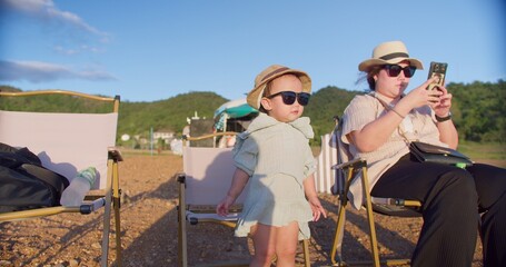 Fototapeta premium A young toddler girl enjoys a sunny day outdoors while her mother films her with a smartphone, capturing precious family moments during a weekend camping trip.