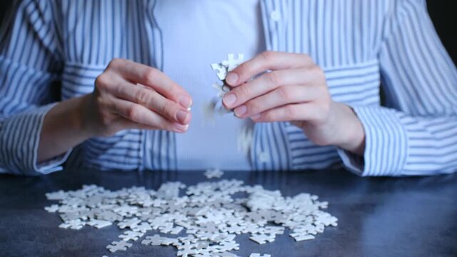 Female hands carefully connecting white jigsaw puzzle pieces together on a dark table, representing problem solving, strategy, and achieving a goal through teamwork and connection