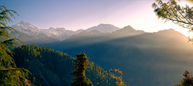 Panoramic Morning Sunrise with Sunbeams over Sainj Valley, Himachal Pradesh Himalayas. the image features dramatic sunbeams and light rays breaking over the Himalayan ridges, illuminating snow-peaks.