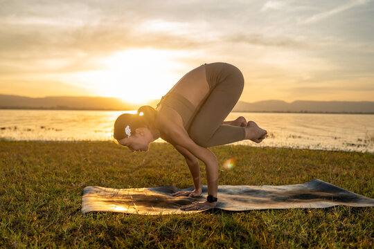 Mindful woman practicing crow pose yoga in scenic sunrise landscape near lake, expressing concentration, strength, flexibility, and peaceful outdoor wellness lifestyle concept.