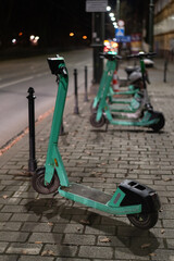 Green electric scooters stand parked on a paved sidewalk along a dark street at night. Modern urban transport rental represents concept of eco friendly micro mobility and convenient commuting. © Tetiana