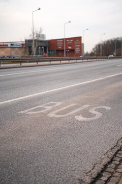 Vertical shot of a dedicated bus lane with white painted text on asphalt road surface. Urban street scene featuring a blurred shopping mall and city lamps in background.